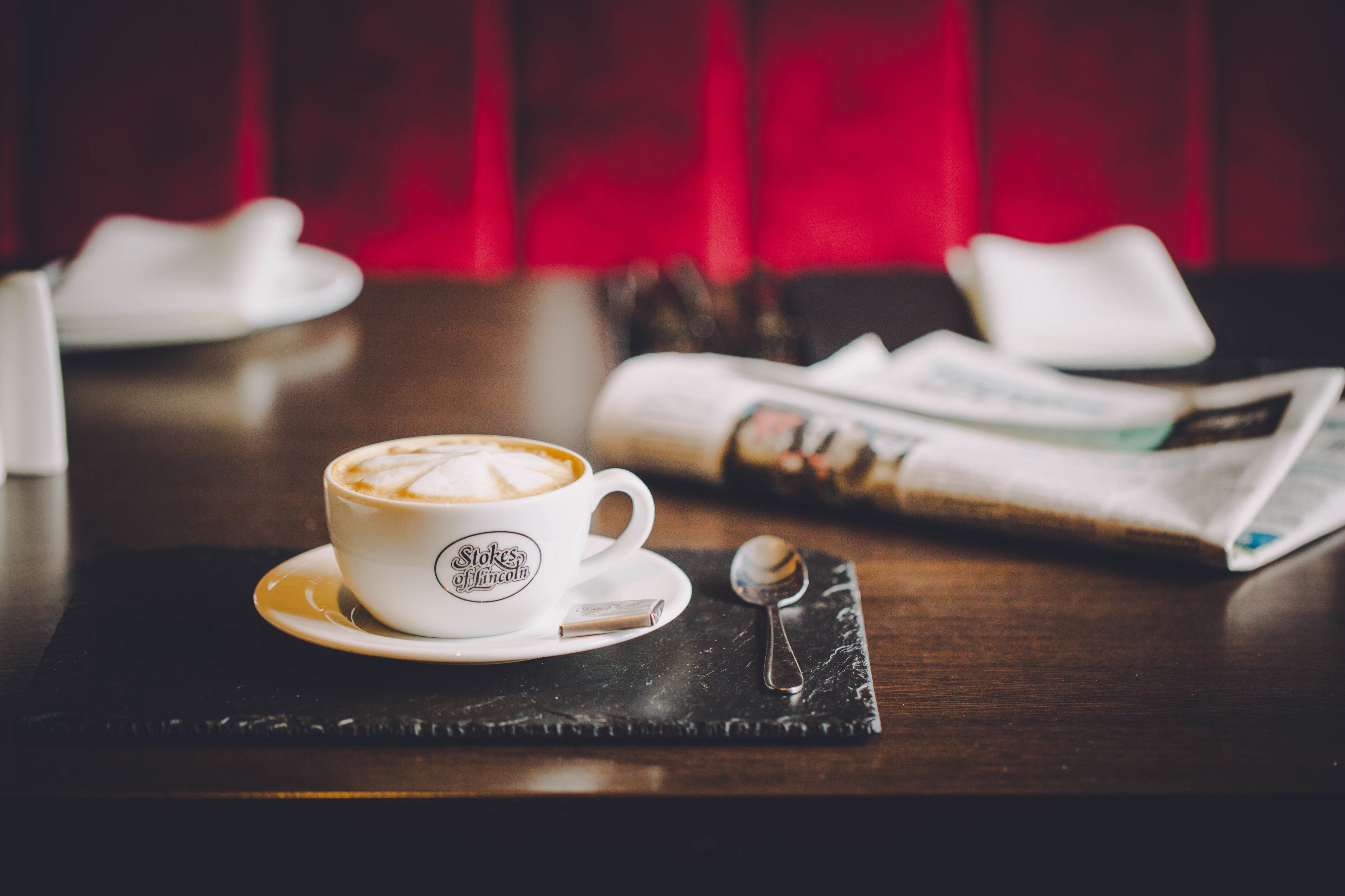 A cup of coffee on a saucer with a spoon beside it, a newspaper, and a napkin on a dark placemat.