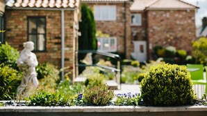 Un jardín con una estatua, un seto bien cuidado y una casa de ladrillo con tejado de tejas.