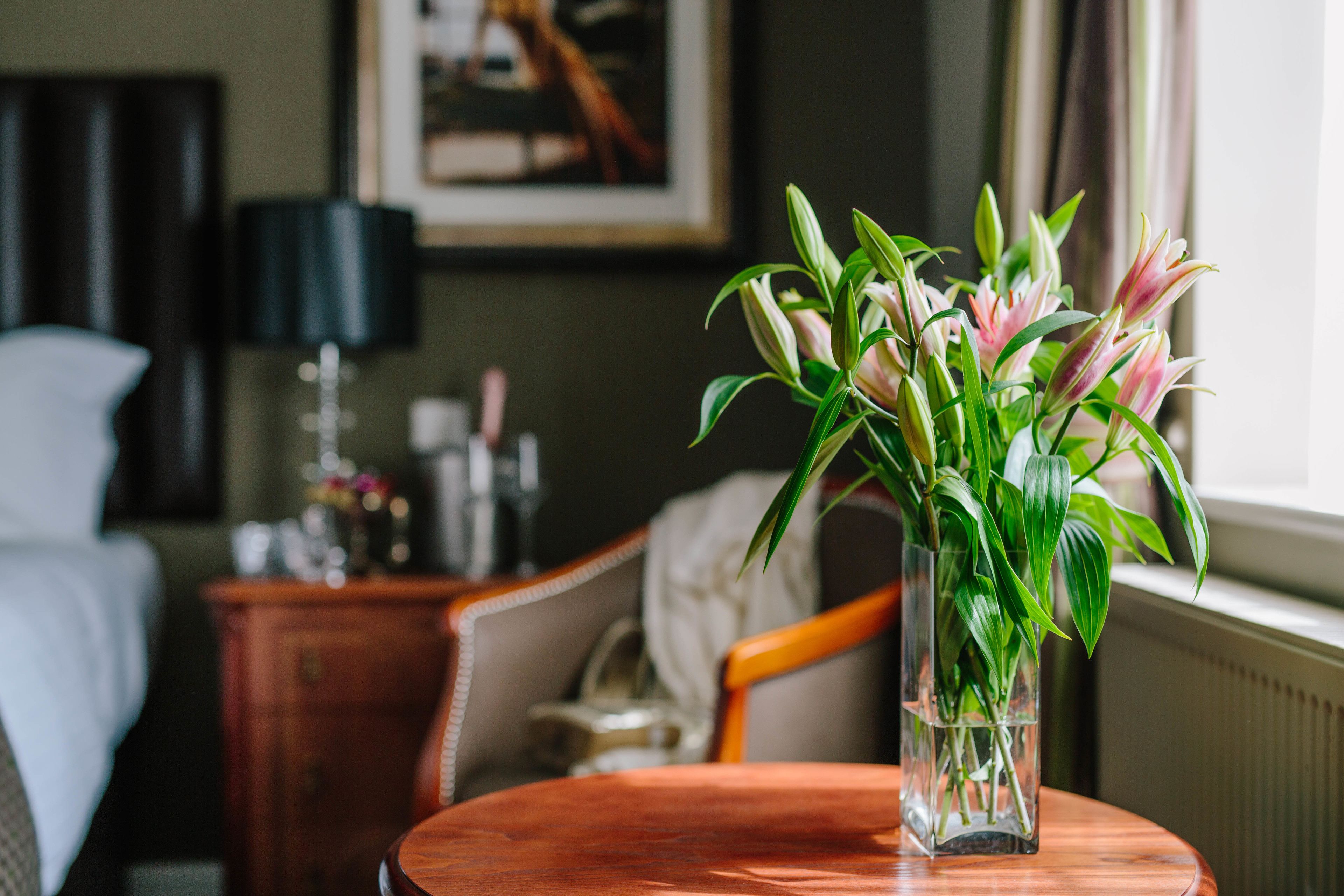 Una habitación de hotel con una mesa de madera, un florero con flores, una cama, una mesita de noche y una lámpara.