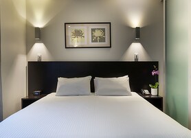 Una camera d'hotel con un letto grande, due comodini, un tavolino da notte, un'opera d'arte incorniciata sopra il letto e una pianta in vaso sul tavolino.