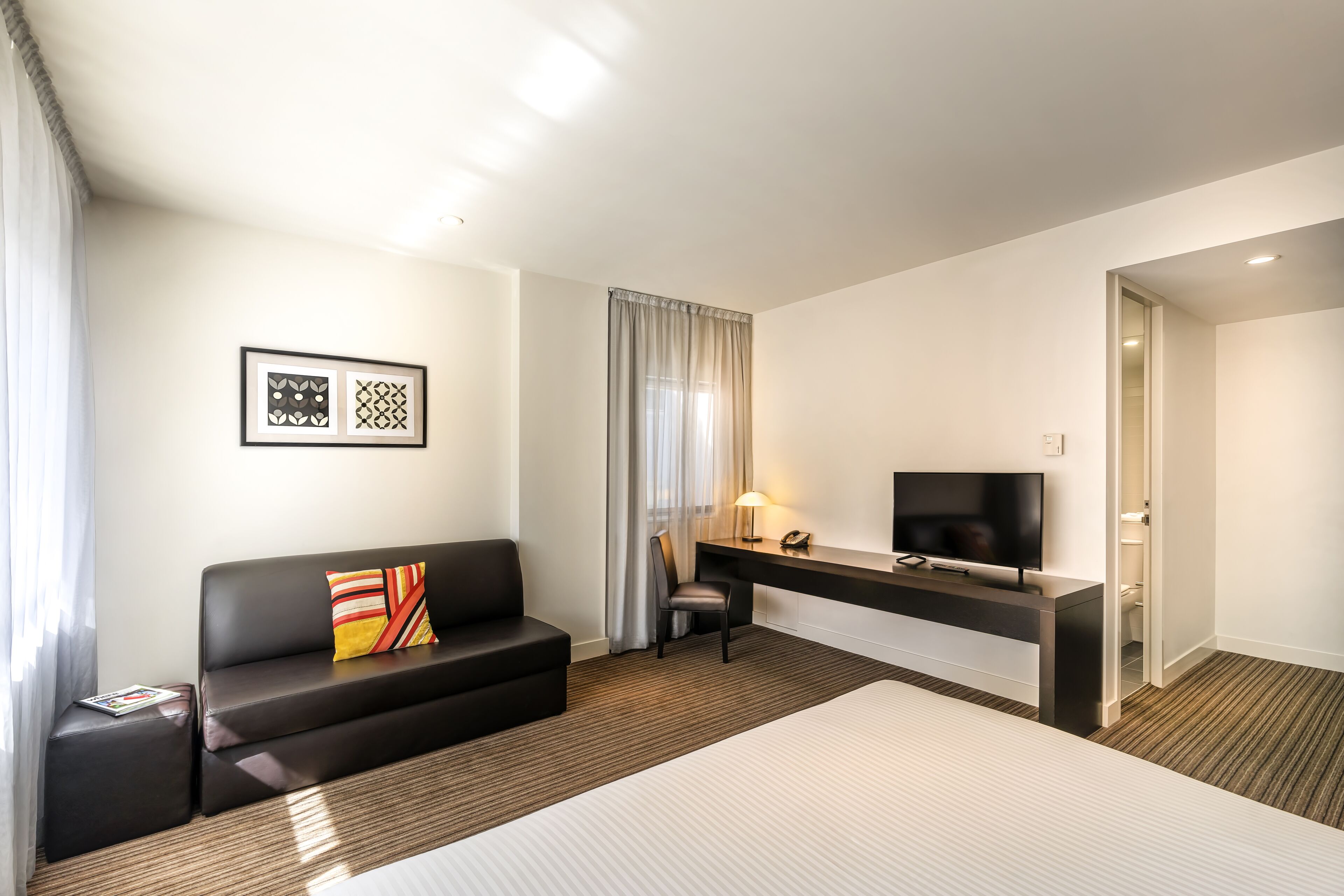 A hotel room with a bed, a black leather sofa, a desk with a TV, and a chair.