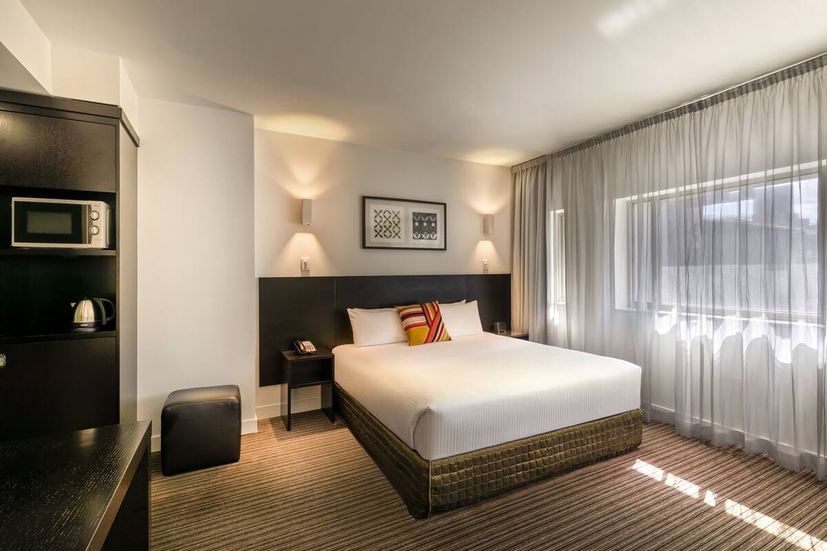 Une chambre d’hôtel avec un grand lit, un bureau, un four à micro-ondes, une table de chevet et une fenêtre avec des rideaux légers.