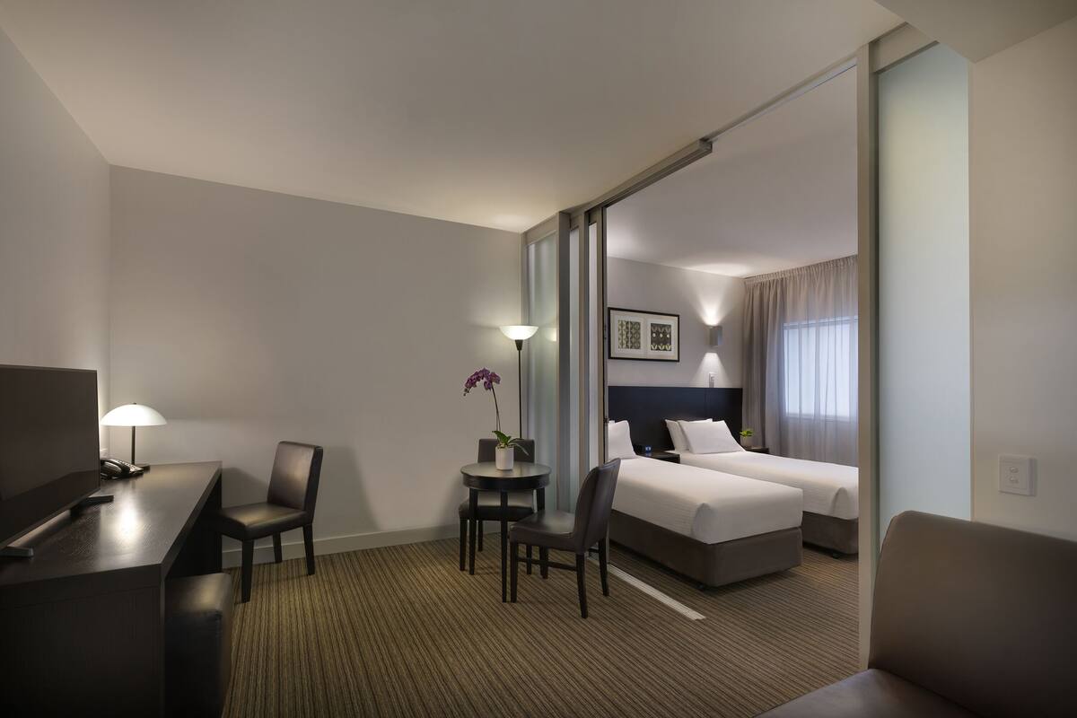 Une chambre d’hôtel équipée d’un bureau, de deux chaises, d’une petite table avec une lampe et une orchidée, d’un lit avec deux oreillers et d’une grande fenêtre avec des rideaux.