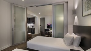 A modern hotel room with a large bed, a separate seating area, and a kitchenette in the background. - Causeway 353 Hotel (Melbourne)