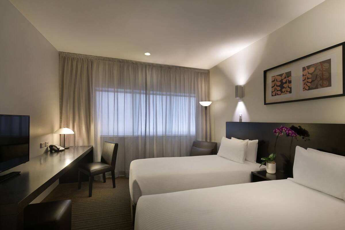 Une chambre d’hôtel avec deux lits, un bureau avec une chaise, une télévision et une fenêtre avec des rideaux.