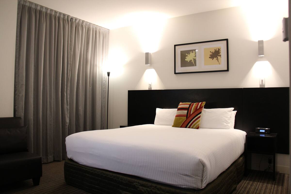 Une chambre d’hôtel avec un grand lit, une table de chevet, une chaise et une fenêtre avec des rideaux.