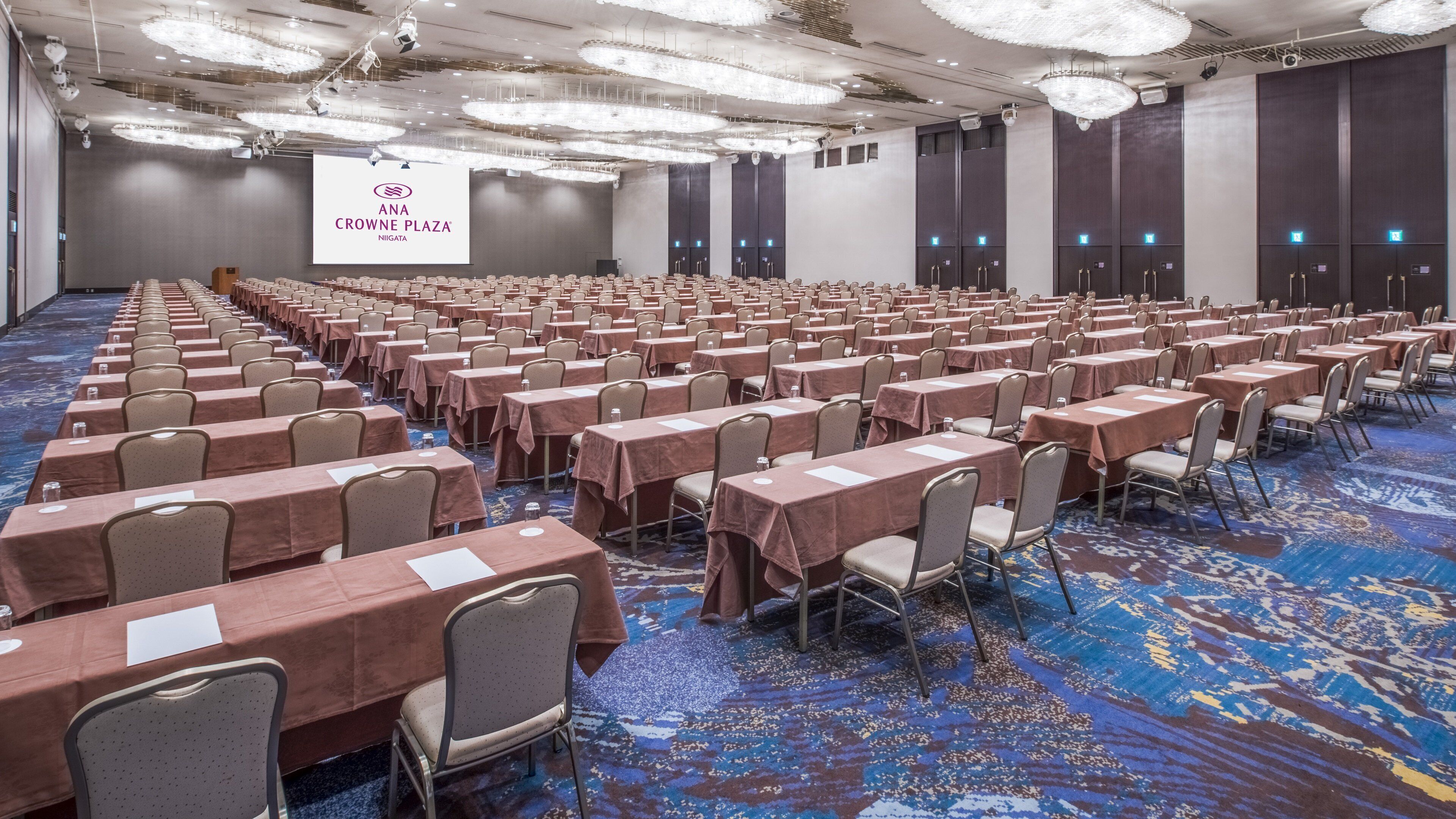 A conference room with rows of tables and chairs, a large screen displaying 'ANA Crowne Plaza Niigata,' and a carpeted floor.