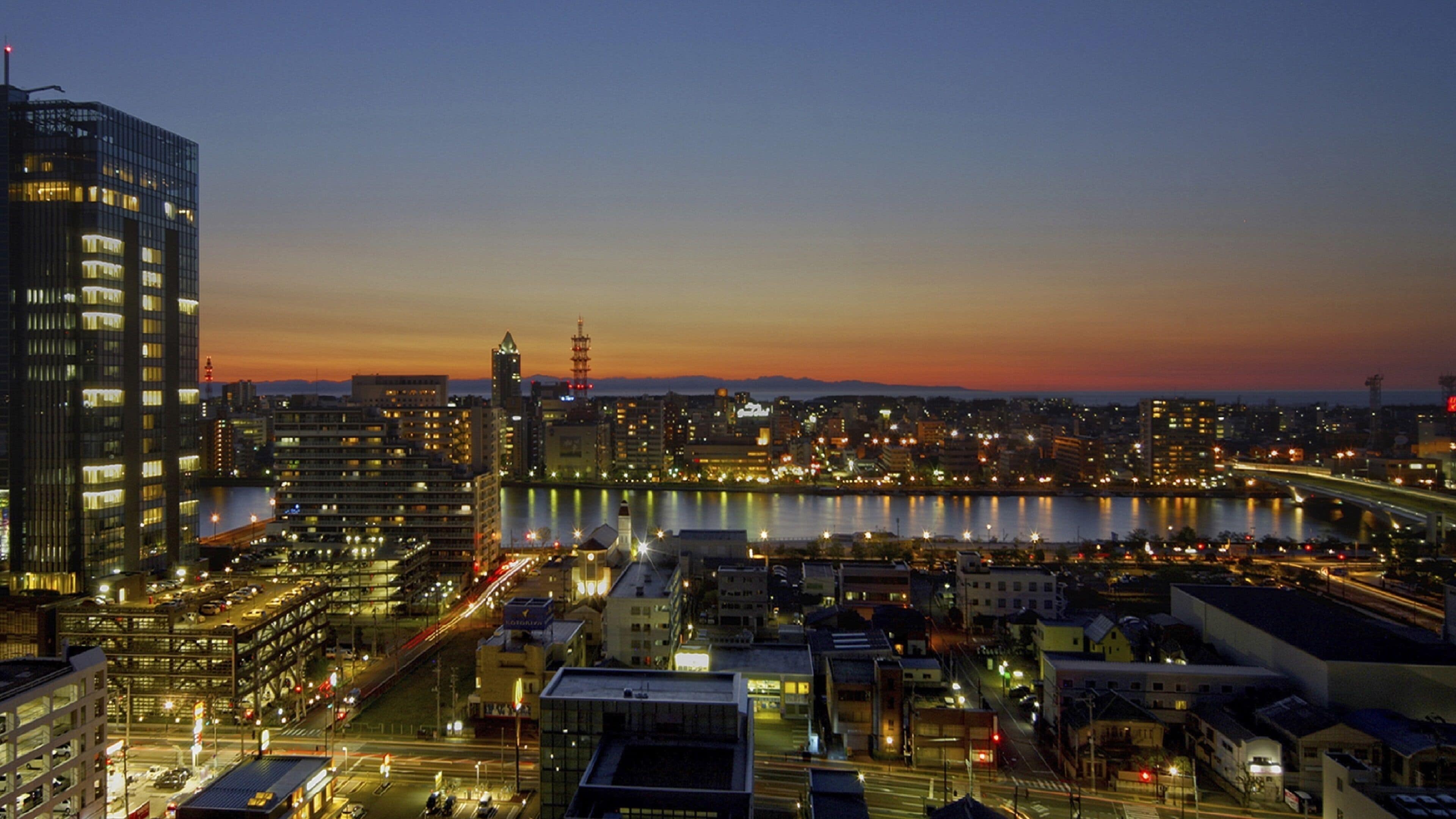 A cityscape at dusk with illuminated buildings, a river, and a bridge.