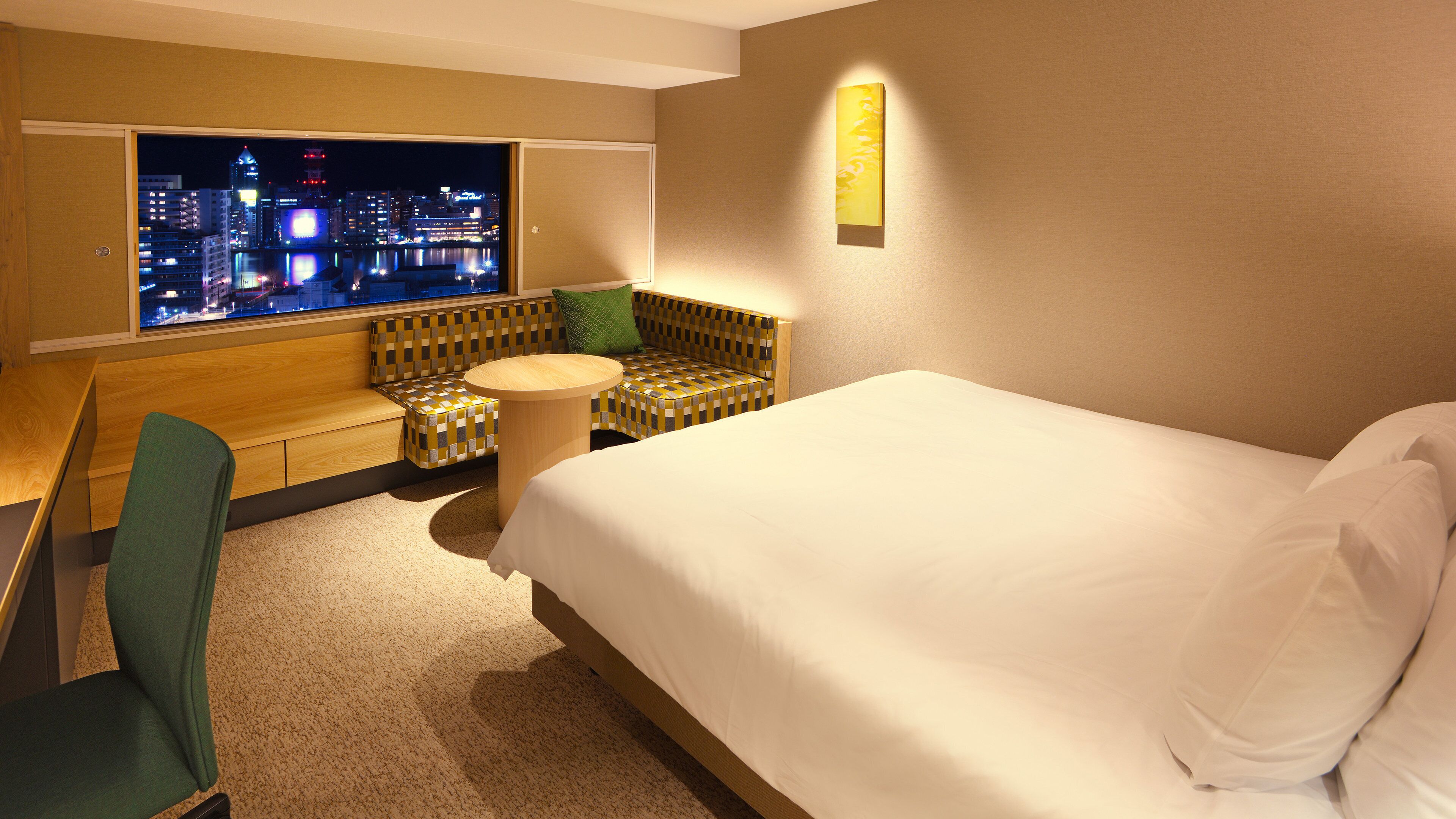 Ein Hotelzimmer mit Bett, einer Sitzecke mit Blick auf die beleuchtete Stadt und einem kleinen runden Tisch.