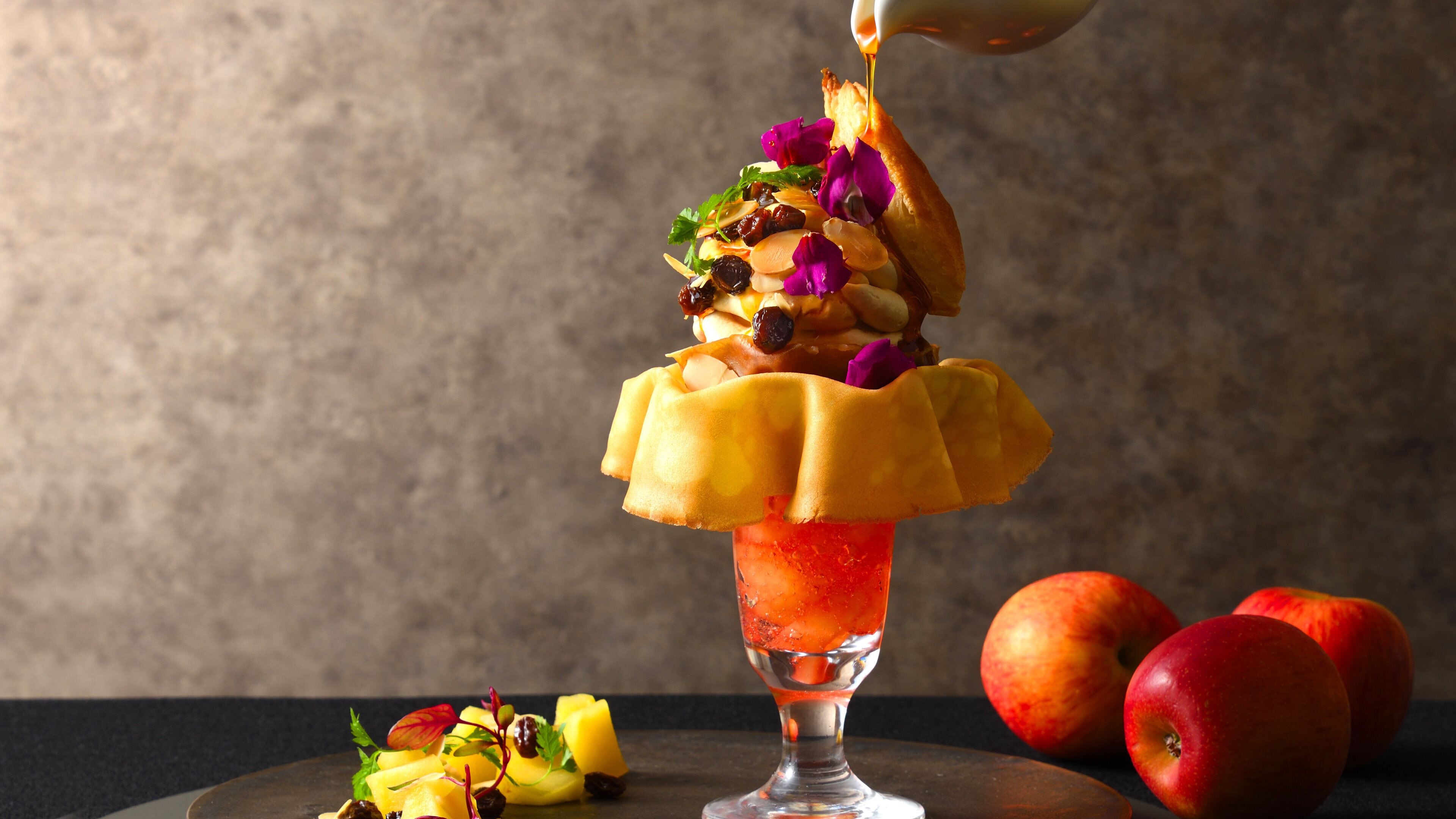 Un postre con borde dorado, cubierto de frutas y nueces variadas, y salsa vertida sobre él.
