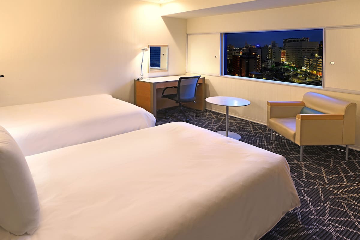 Ein Hotelzimmer mit zwei Betten, einem Schreibtisch, einem Stuhl und Blick auf die beleuchtete Stadt.