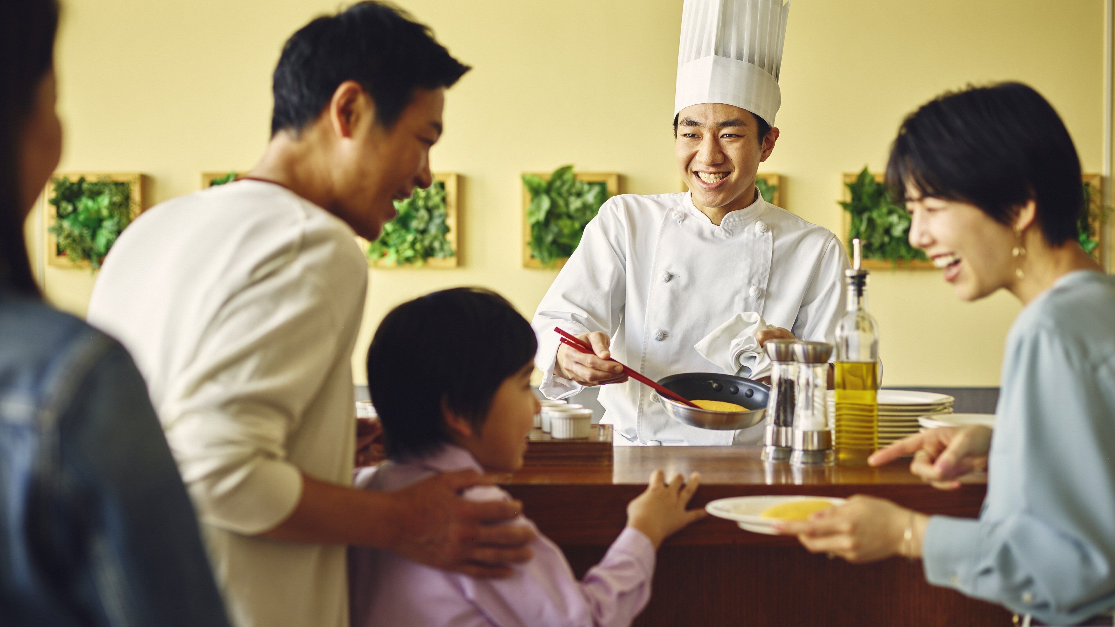 A chef in a white uniform cooking at a counter with a family nearby.