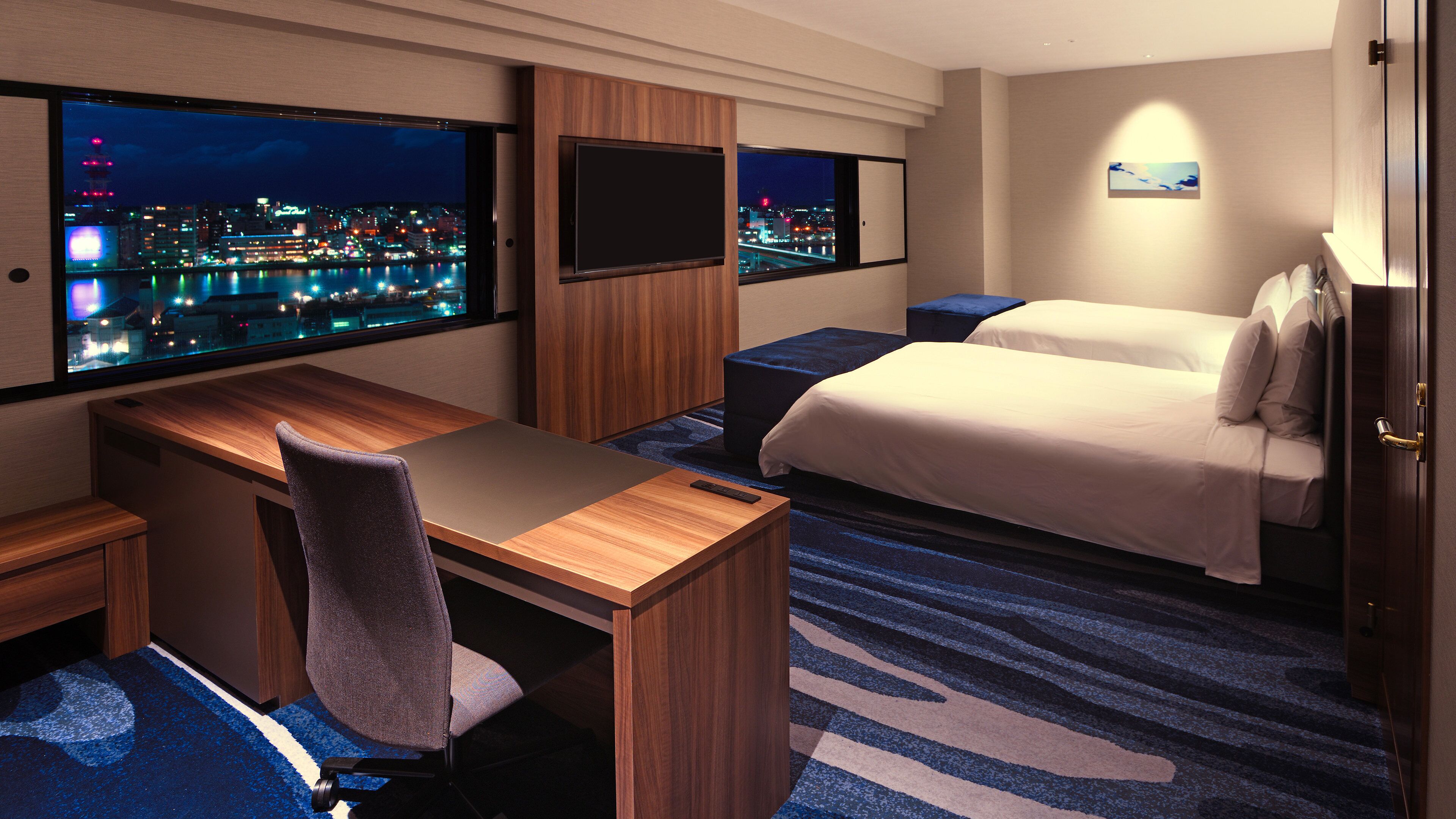 A hotel room with a desk, chair, two beds, and a large window with a city view.