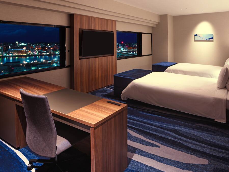Een hotelkamer met een bureau, stoel, twee bedden en een groot raam met uitzicht op de stad.