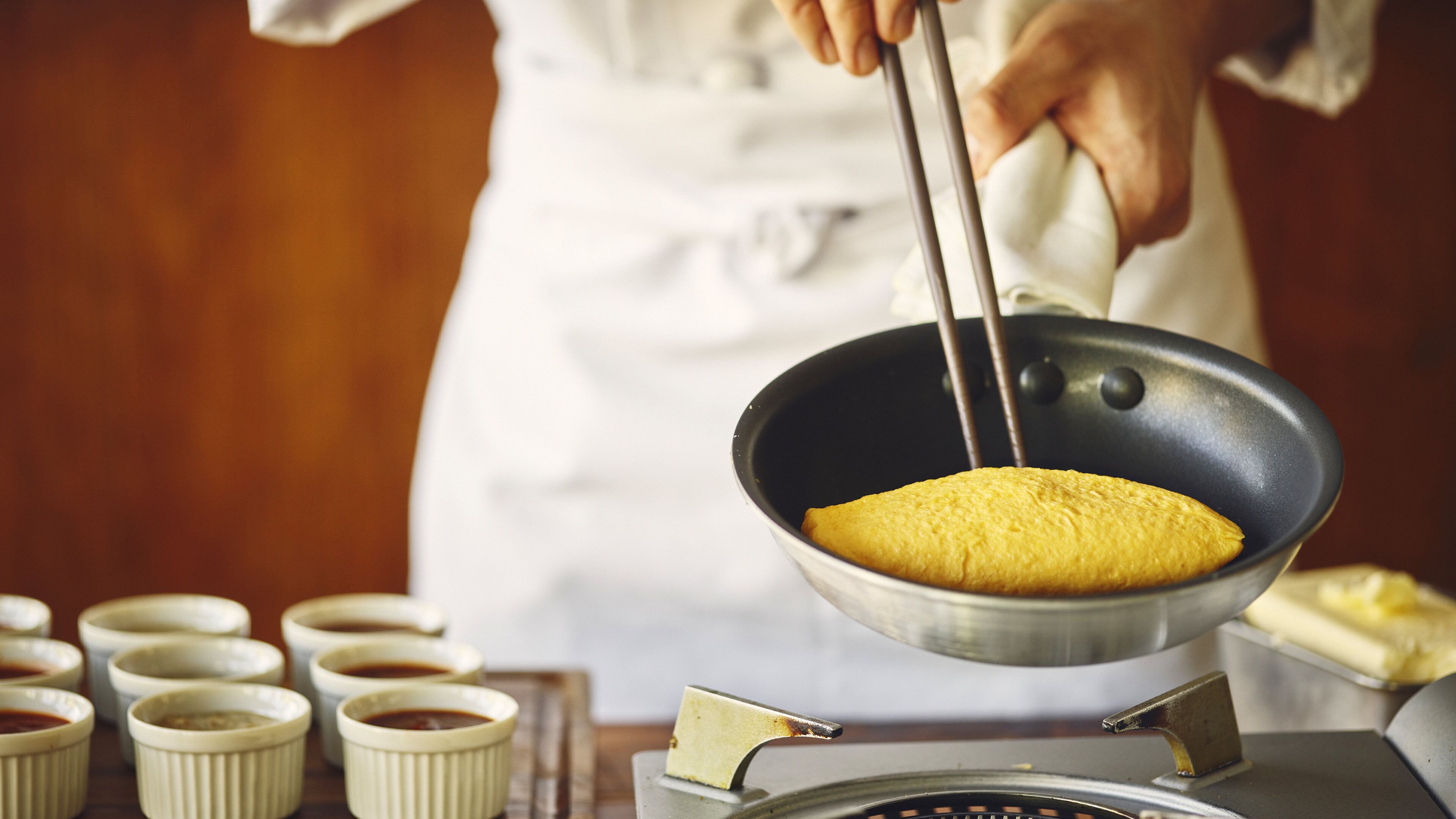 Una persona cocinando en una cocina con una sartén, una estufa y varios recipientes pequeños.