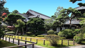 Ein traditionelles japanisches Gebäude mit einem Ziegeldach, umgeben von gepflegter Bepflanzung und einem Holzzaun.