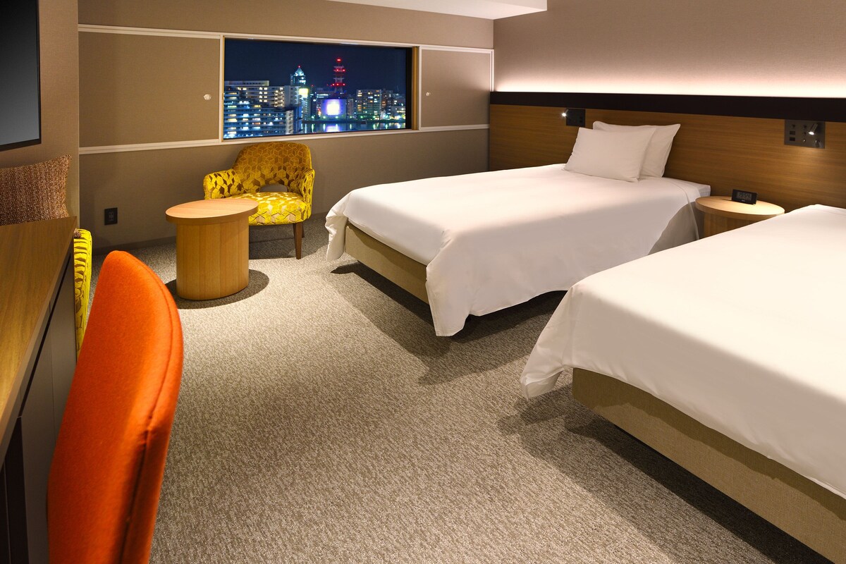 Ein Hotelzimmer mit zwei Betten, einem kleinen runden Tisch, einem gelben Sessel und Blick auf die Stadt durch das Fenster.