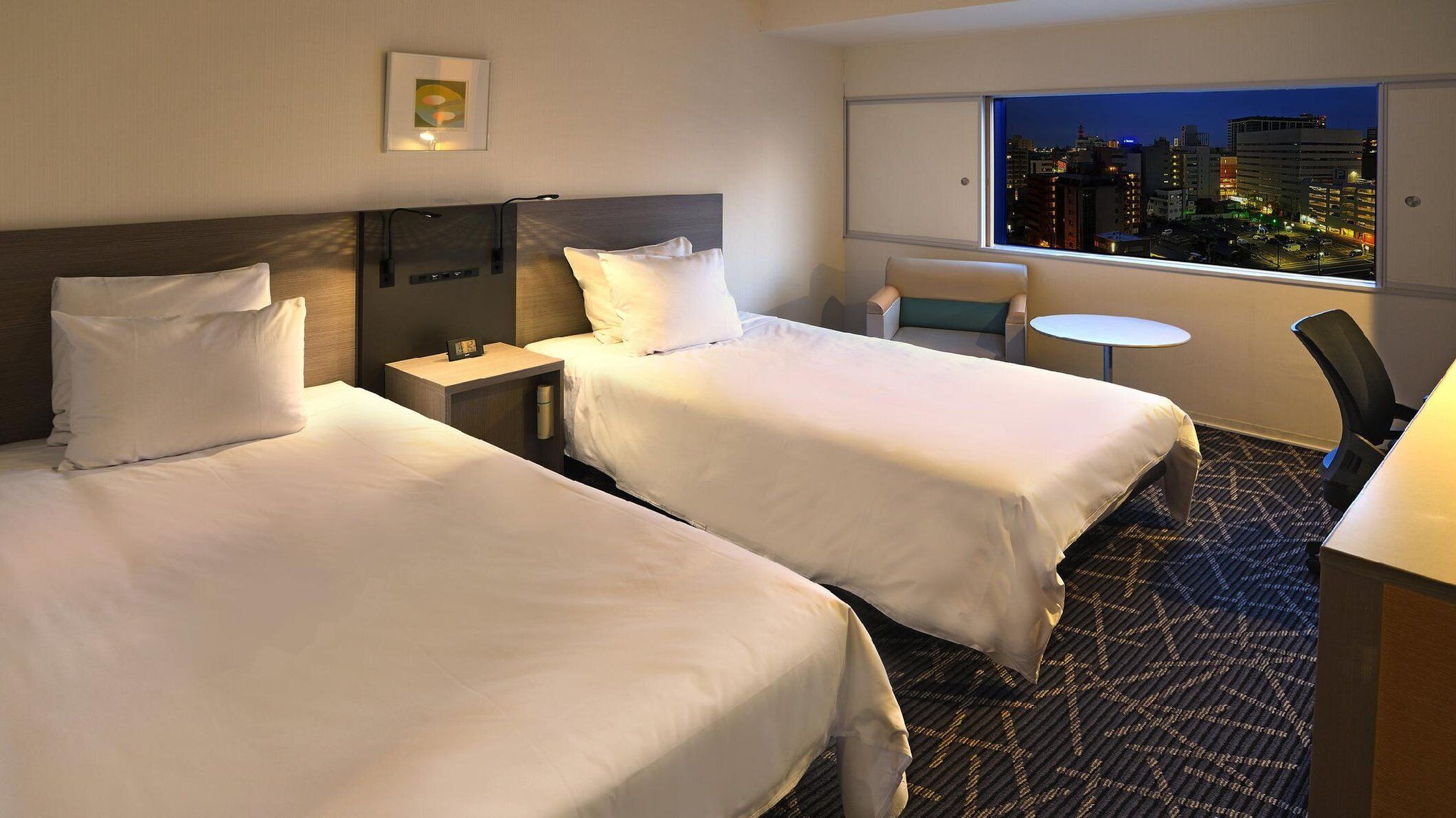 A hotel room with two beds, a small table, a chair, and a view of a cityscape at night.