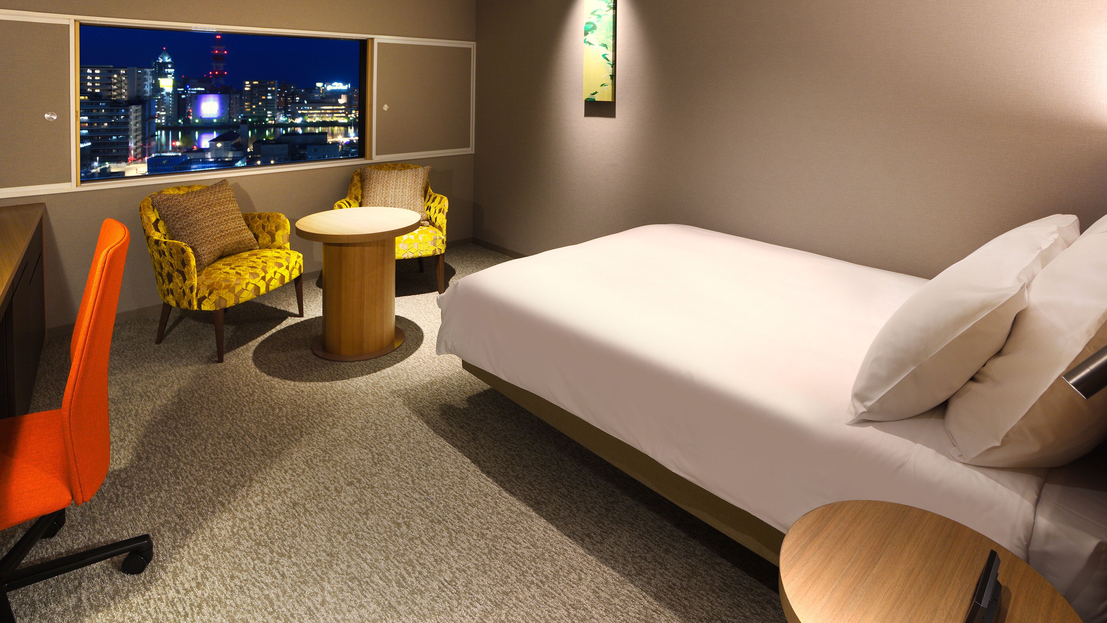 Ein Hotelzimmer mit einem Bett, zwei gelben Sessel, einem kleinen runden Tisch, Blick auf eine nächtliche Skyline und einer Wandleuchte.