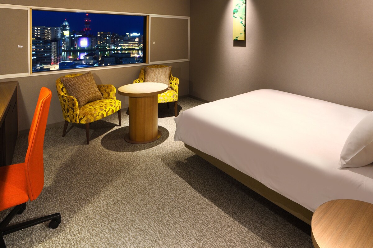 Ein Hotelzimmer mit einem Bett, zwei gelben Sessel, einem kleinen runden Tisch, Blick auf eine nächtliche Skyline und einer Wandleuchte.