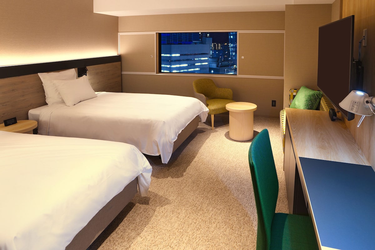 Une chambre d’hôtel avec deux lits, une petite table ronde, une chaise, un bureau et une grande fenêtre donnant sur la ville.