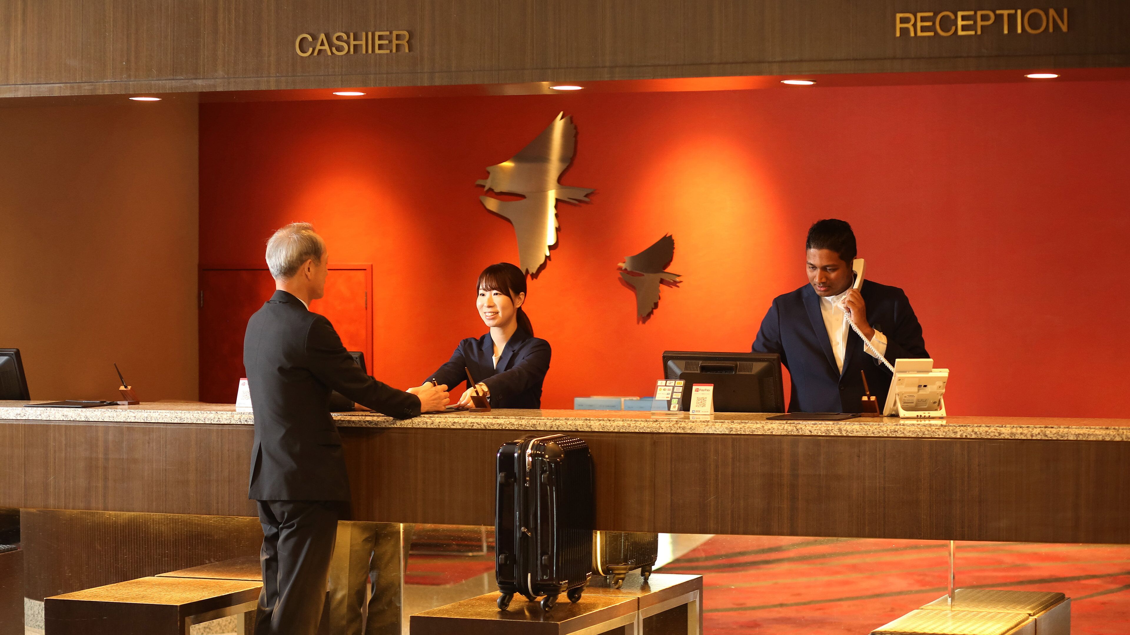 A hotel reception area with a counter, a cash register, and a telephone. There are three people present: a receptionist, a guest, and a hotel staff member. The reception area has a red wall with a bird mural.