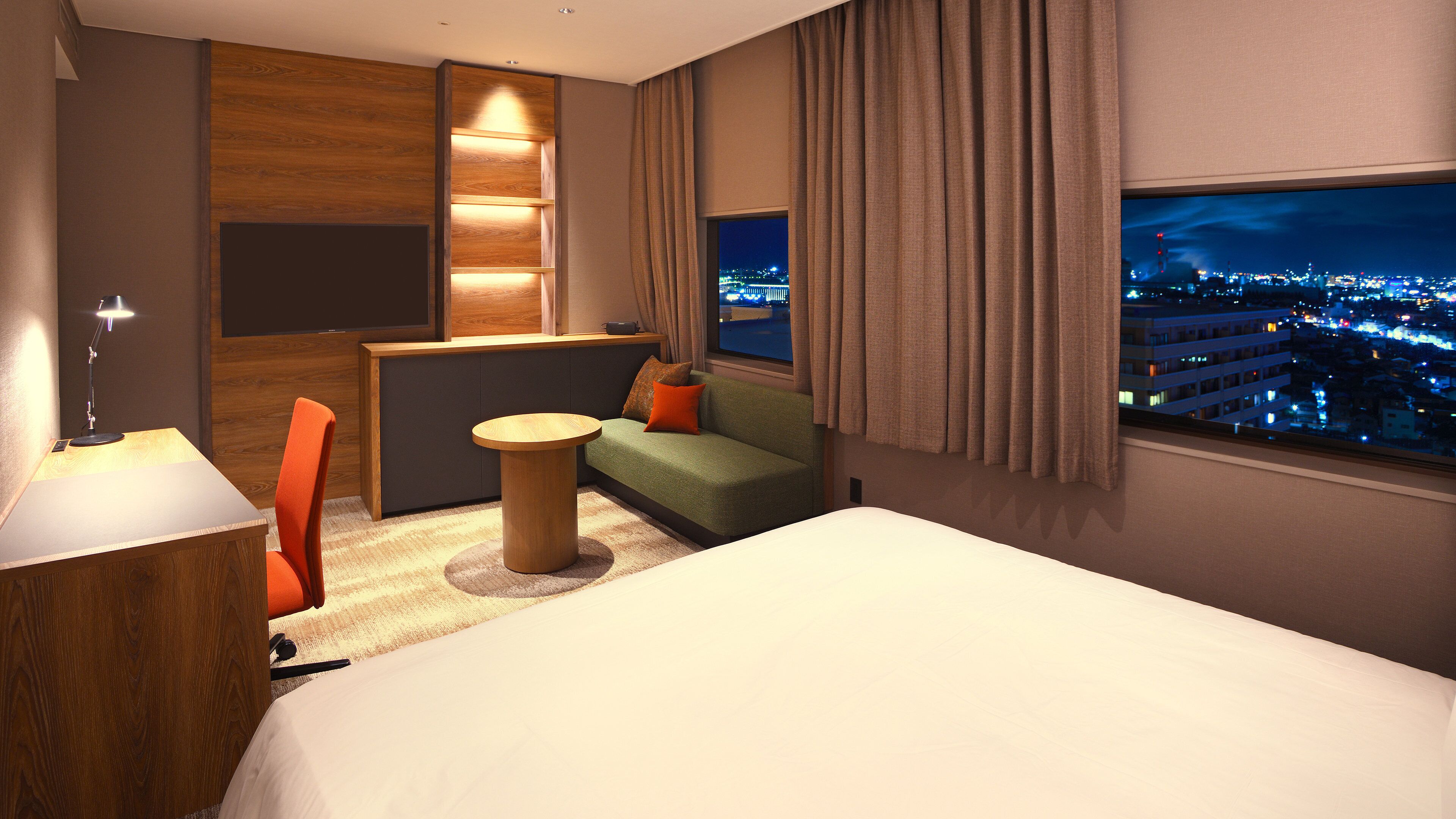 A hotel room with a bed, a sofa, a small table, a desk with a lamp, a window with a city view, and a wooden panel wall.