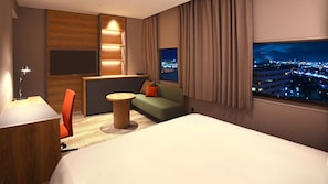 A hotel room with a bed, a sofa, a small table, a desk with a lamp, a window with a city view, and a wooden panel wall.