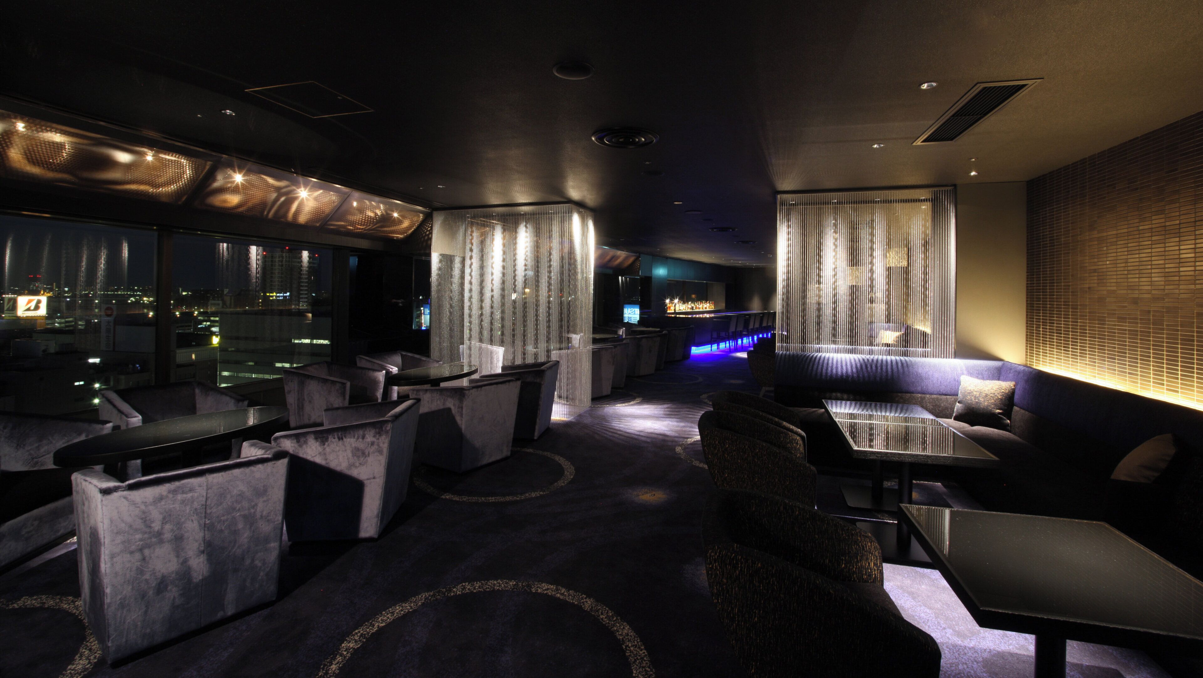 A modern lounge area with seating and a bar, featuring a city view through large windows.