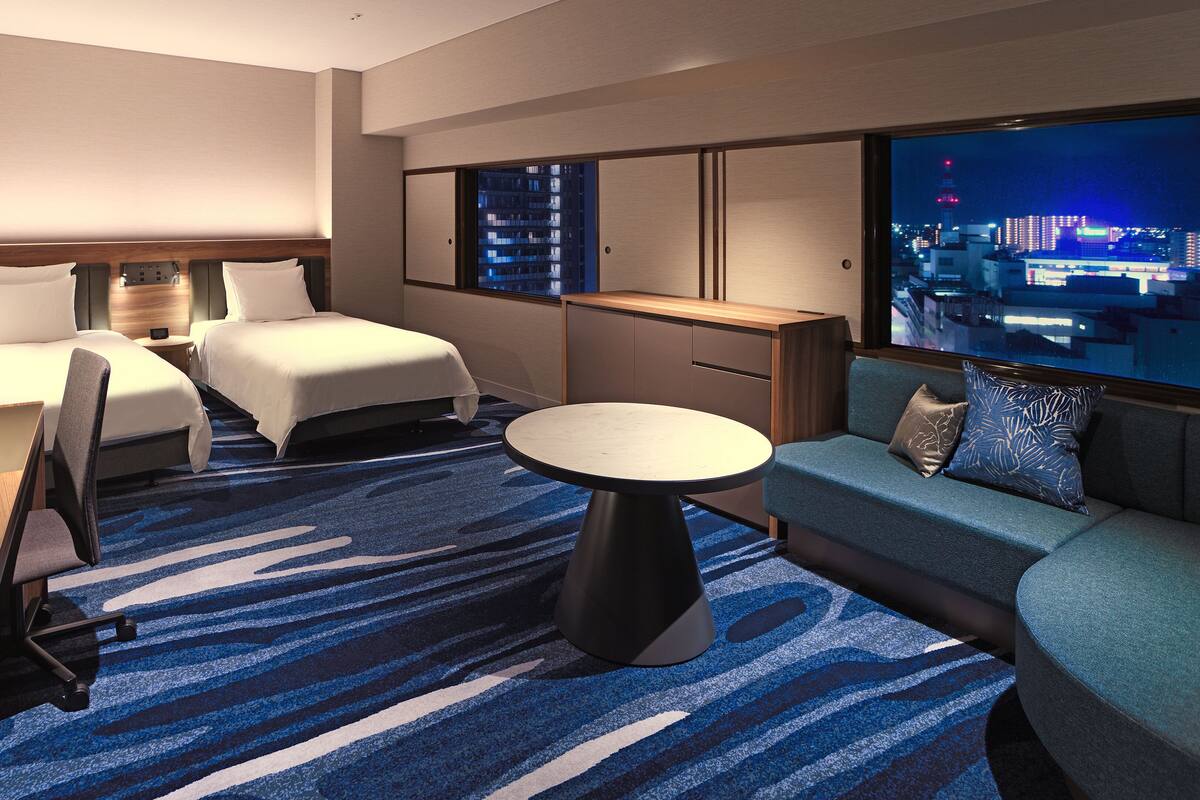 Ein Hotelzimmer mit zwei Betten, einem Schlafsofa, einem Tisch und einem Fernseher, der eine Stadtansicht zeigt.