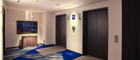 Un pasillo de hotel con alfombra azul y color crema, vista nocturna de la ciudad, un ascensor y una puerta con un letrero azul que indica el número de habitación.