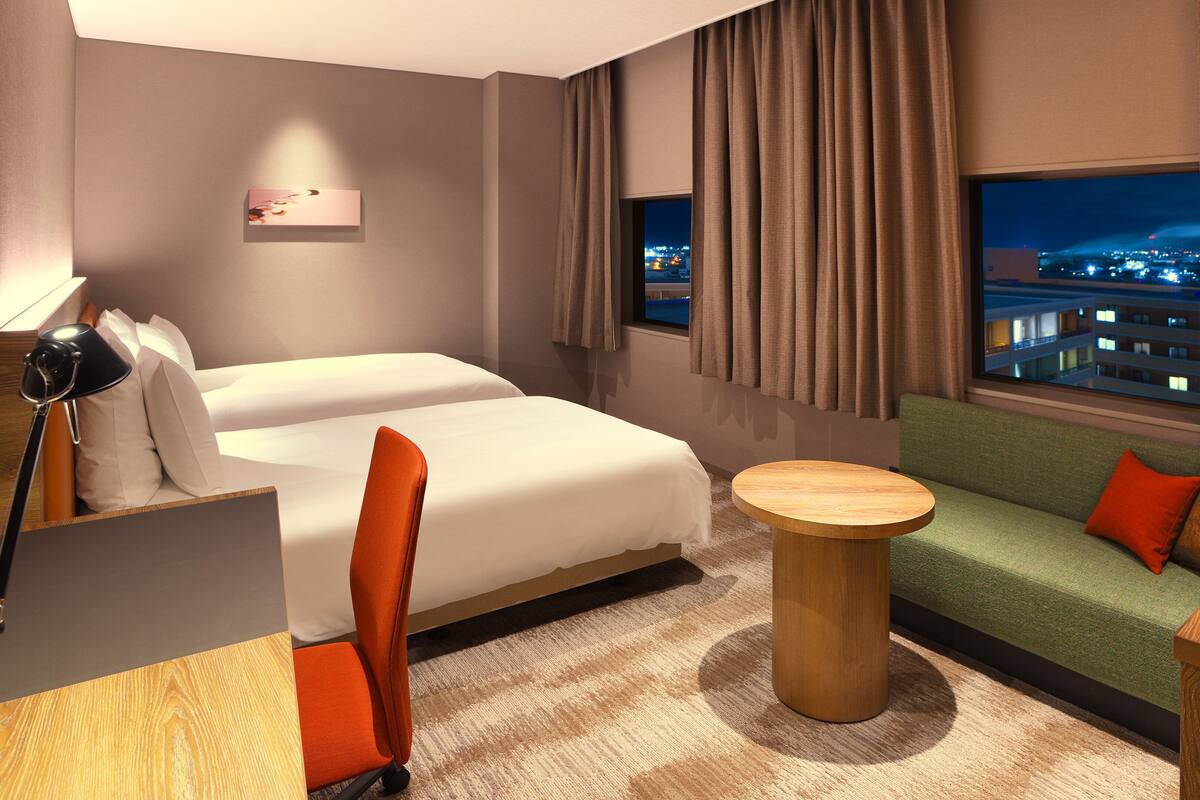 Ein Hotelzimmer mit einem Bett, zwei Nachttischen, einem Sofa, einem kleinen runden Tisch und Blick auf eine nächtliche Stadtansicht.
