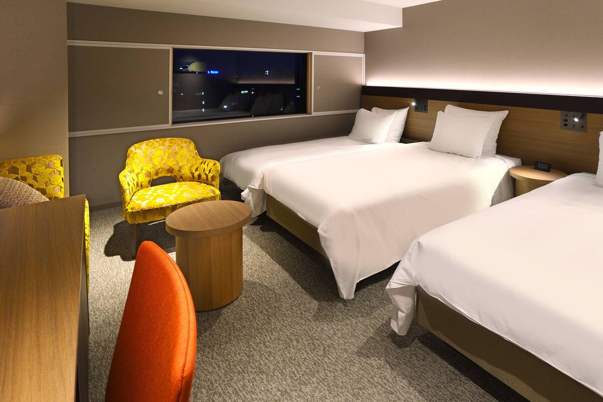 Ein Hotelzimmer mit zwei Betten, einem Fenster mit Blick auf die Stadt, einem gelben Sessel und einem kleinen Holfnachtkästchen.