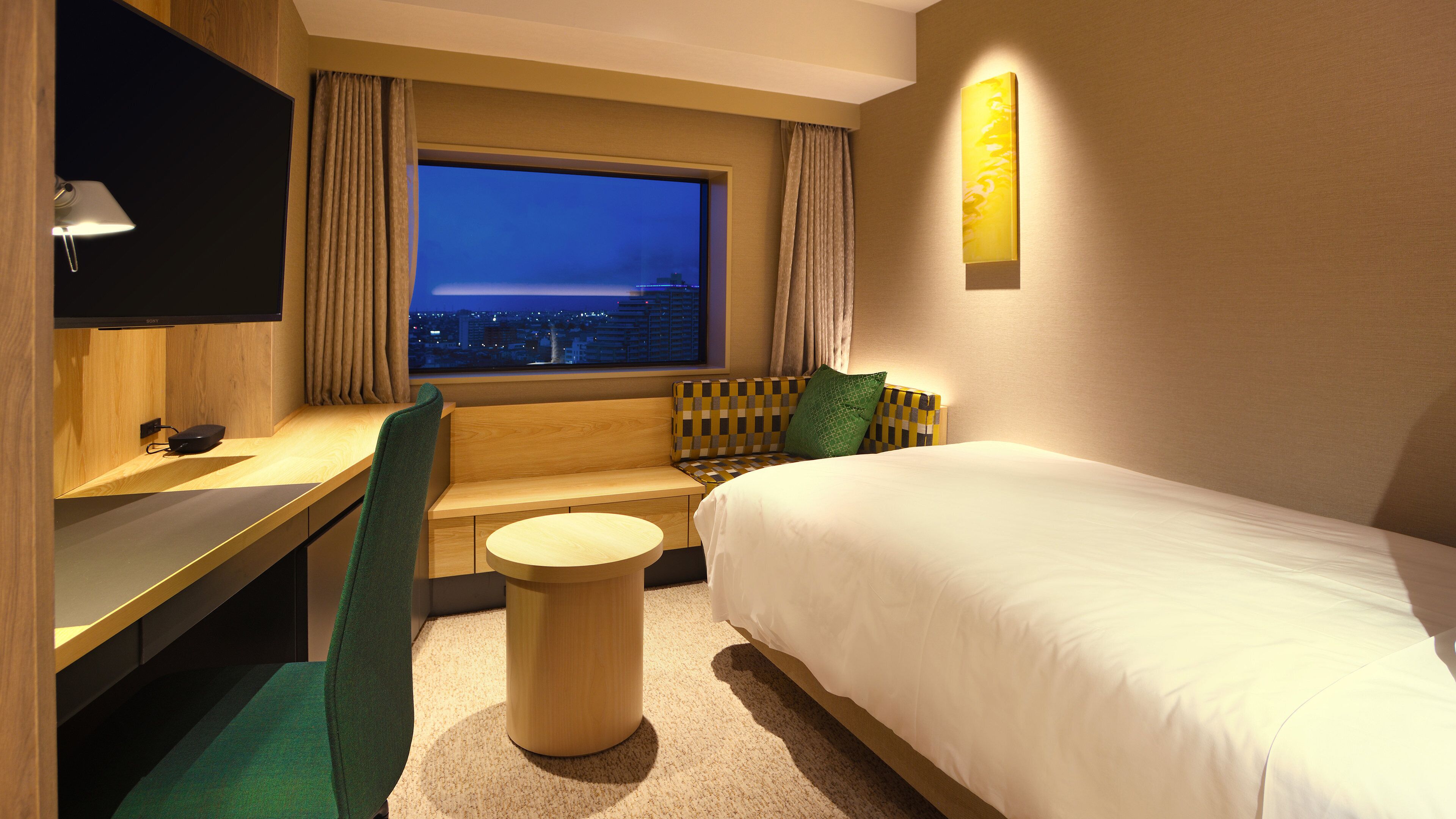 Ein Hotelzimmer mit Bett, Schreibtisch, Stuhl, rundem Tisch, Fernseher und Fenster mit Blick auf die Stadt.