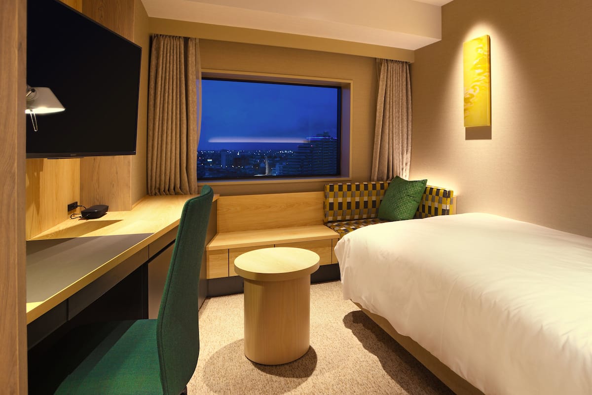 Ein Hotelzimmer mit Bett, Schreibtisch, Stuhl, rundem Tisch, Fernseher und Fenster mit Blick auf die Stadt.