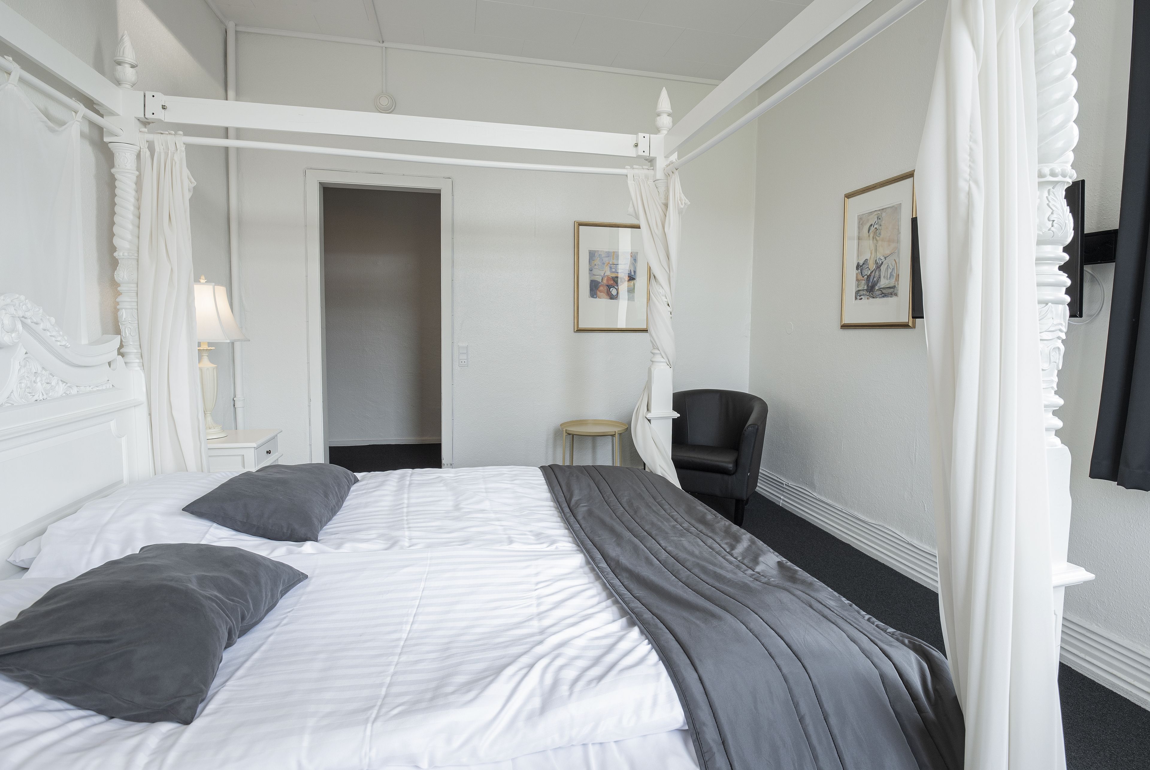 Una cama con dosel, ropa de cama blanca y almohadas grises, un sillón de cuero negro y una pequeña mesa redonda con una lámpara.