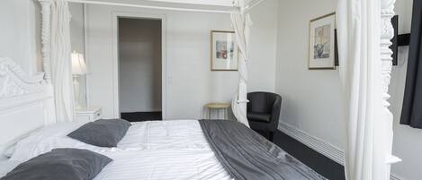 Una cama con dosel, ropa de cama blanca y almohadas grises, un sillón de cuero negro y una pequeña mesa redonda con una lámpara.