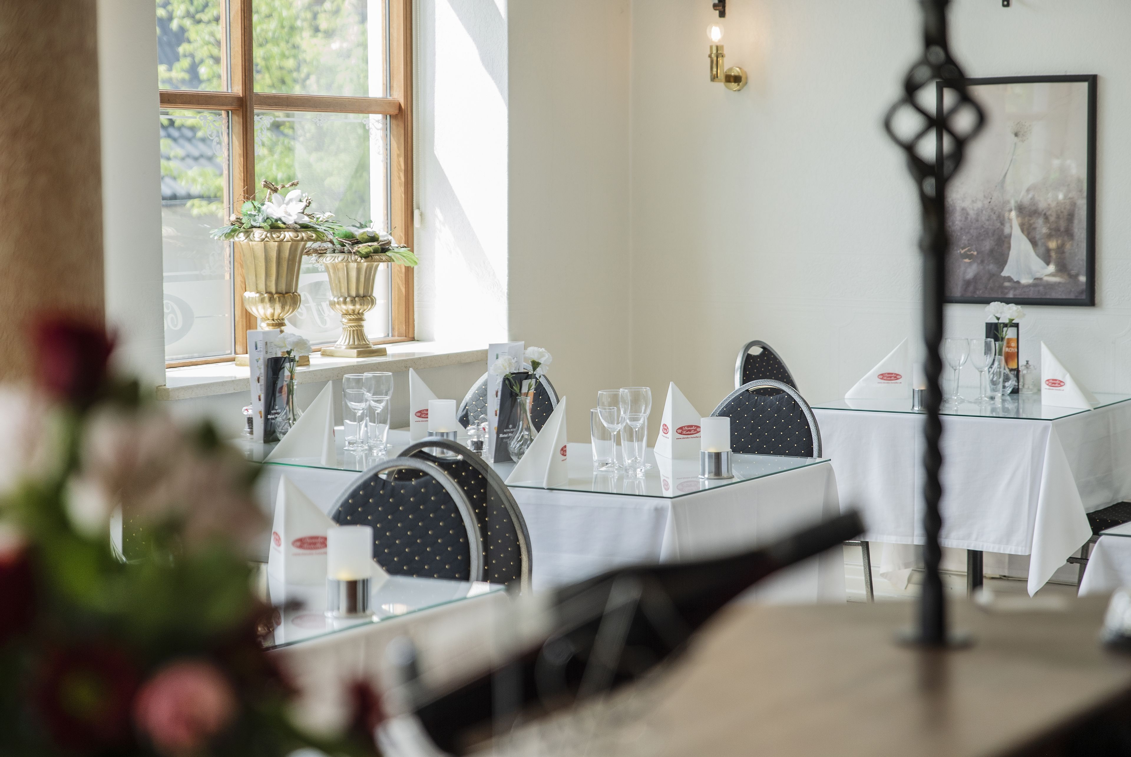 A dining area with tables set for service, a window with potted plants, and a wall-mounted light fixture.
