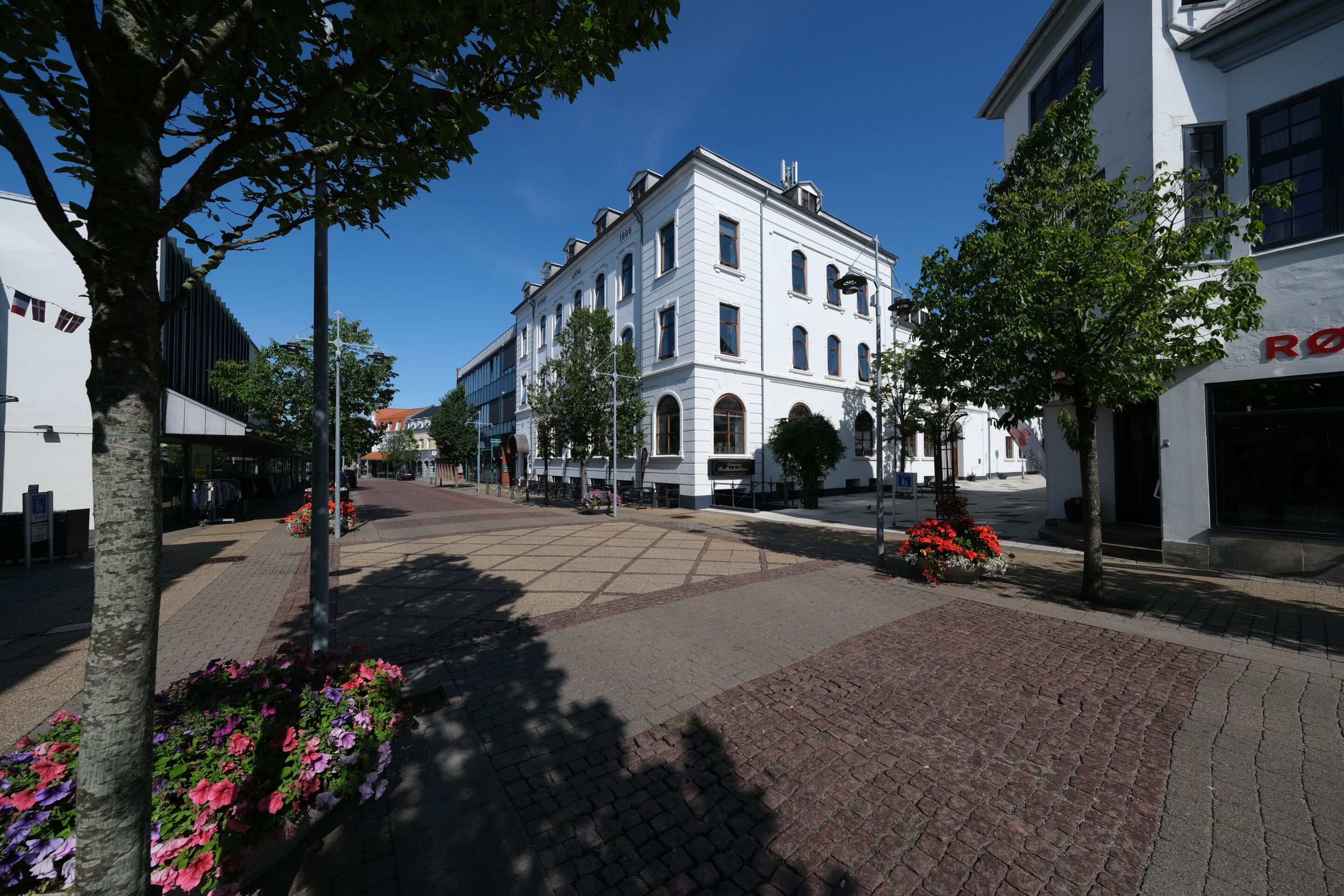 A street with a white building, trees, and flower beds.