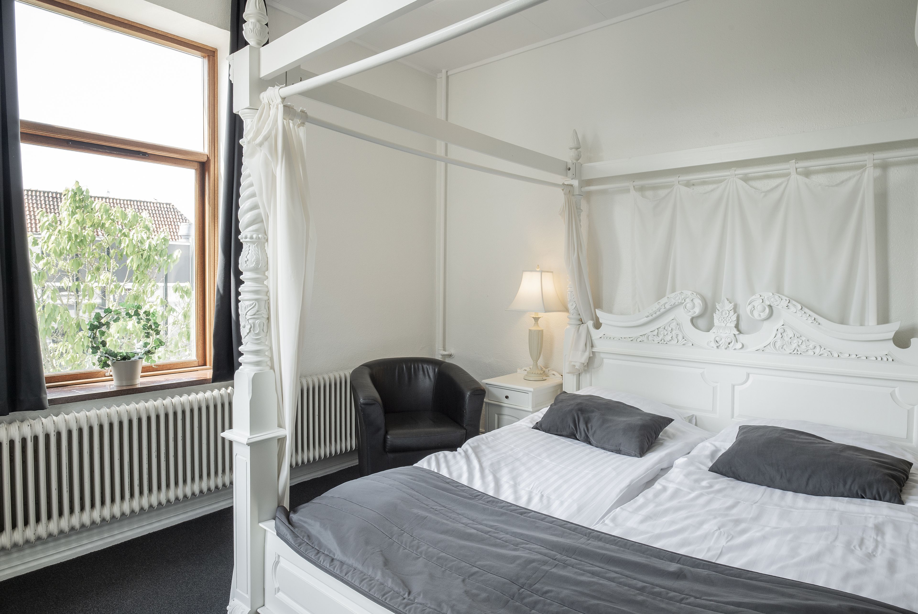 Una cama con dosel con ropa de cama blanca, una silla negra, una mesita de noche con una lámpara y una ventana con vista a un edificio y un árbol.