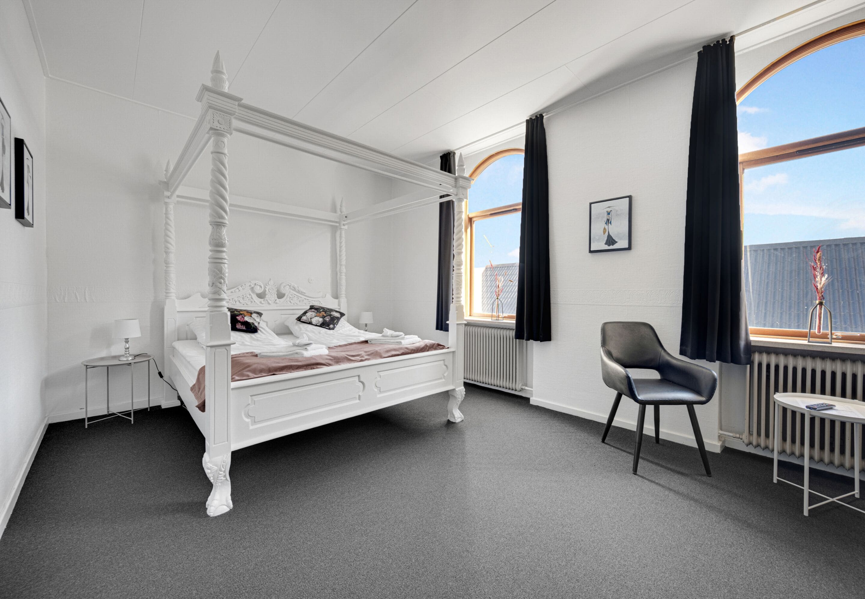 A white four-poster bed with a wooden headboard, a grey upholstered chair, and a small table with a lamp.