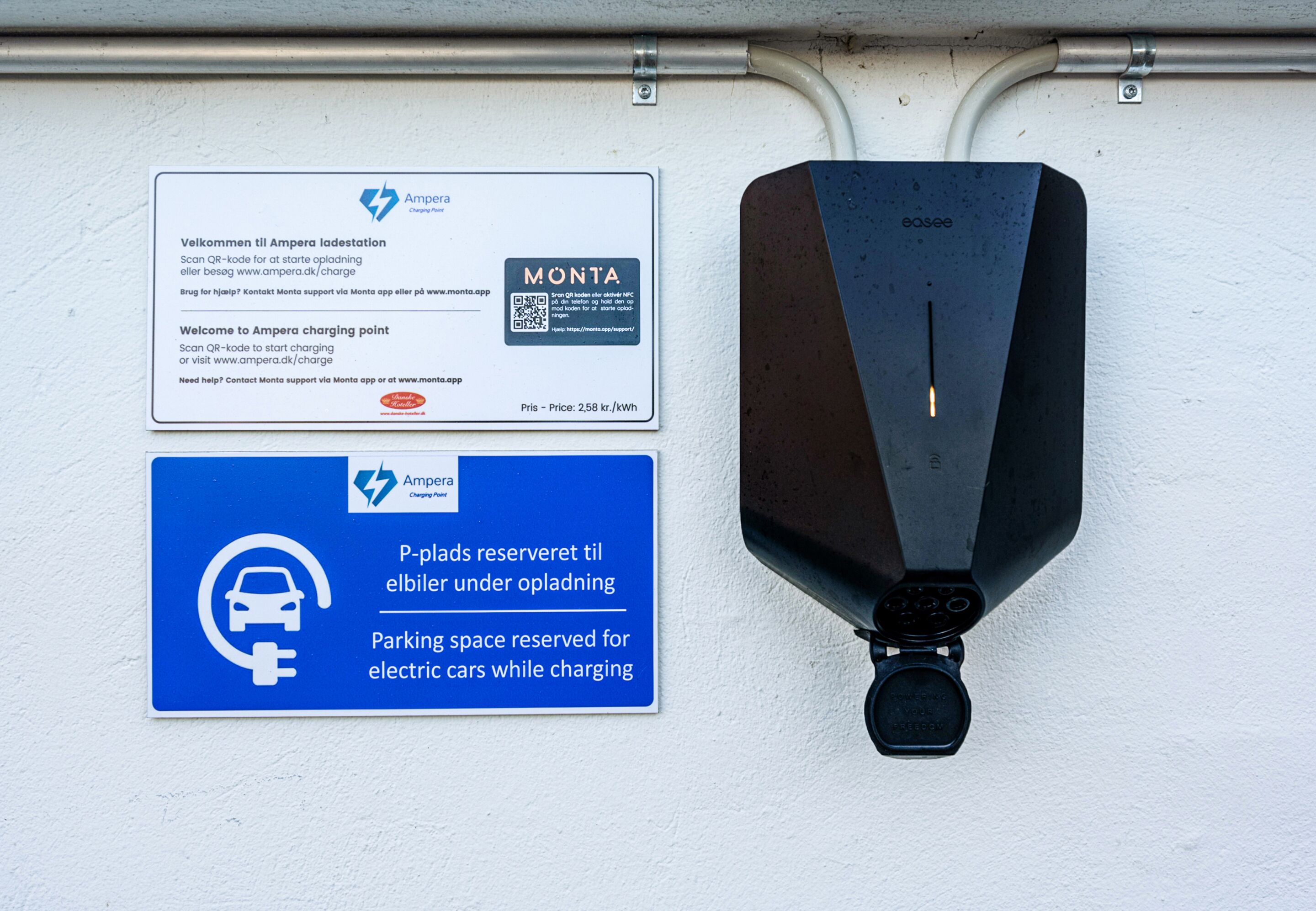 Estación de carga con un cartel que indica punto de carga Ampera, un código QR para iniciar la carga y un letrero azul que reserva plaza de aparcamiento para coches eléctricos durante la carga.