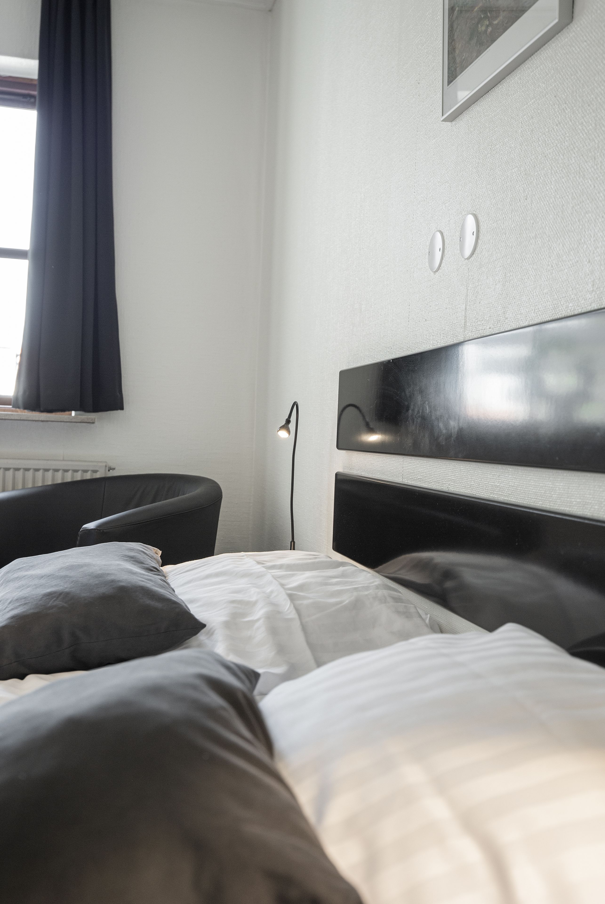 Una habitación de hotel moderna con una cama grande, una mesita de noche, una lámpara y un ventanal con cortinas.