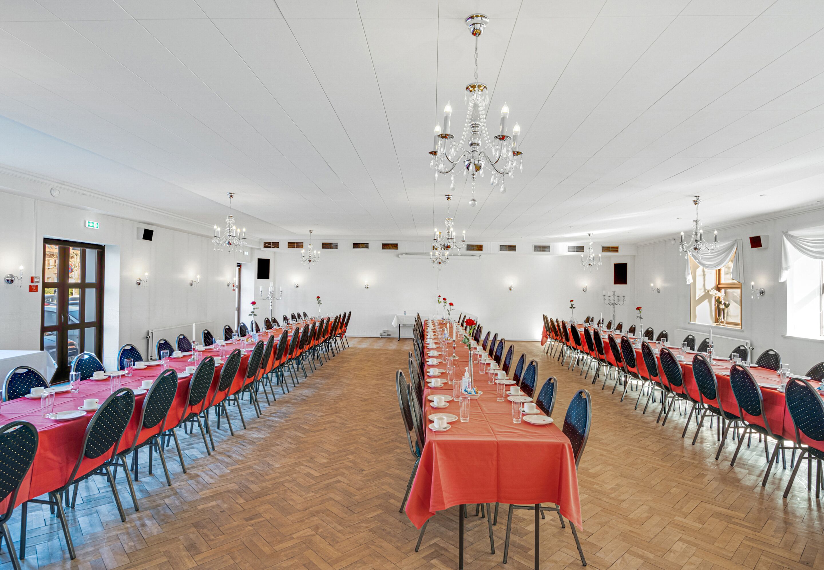 A large conference room with long tables, chairs, chandeliers, and a wooden floor.