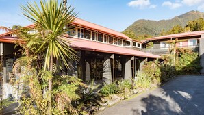 A building with a red roof and multiple windows, surrounded by lush greenery and a mountain in the background. - Heartland Hotel Glacier Country (Fox Glacier)