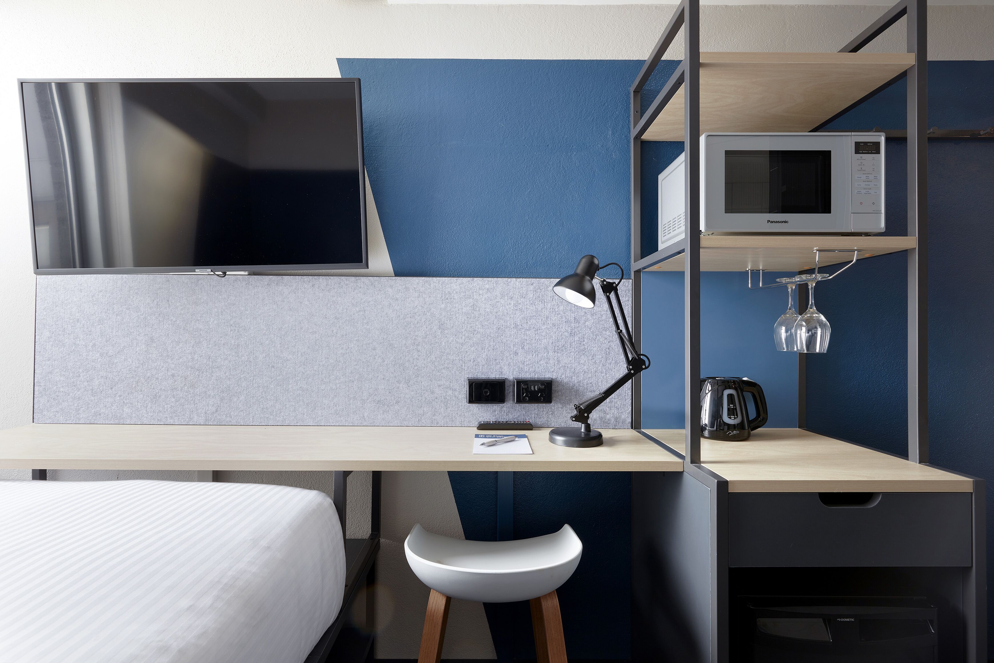 A modern hotel room with a bed, a desk with a computer, a TV, a microwave, a coffee maker, and a small stool.