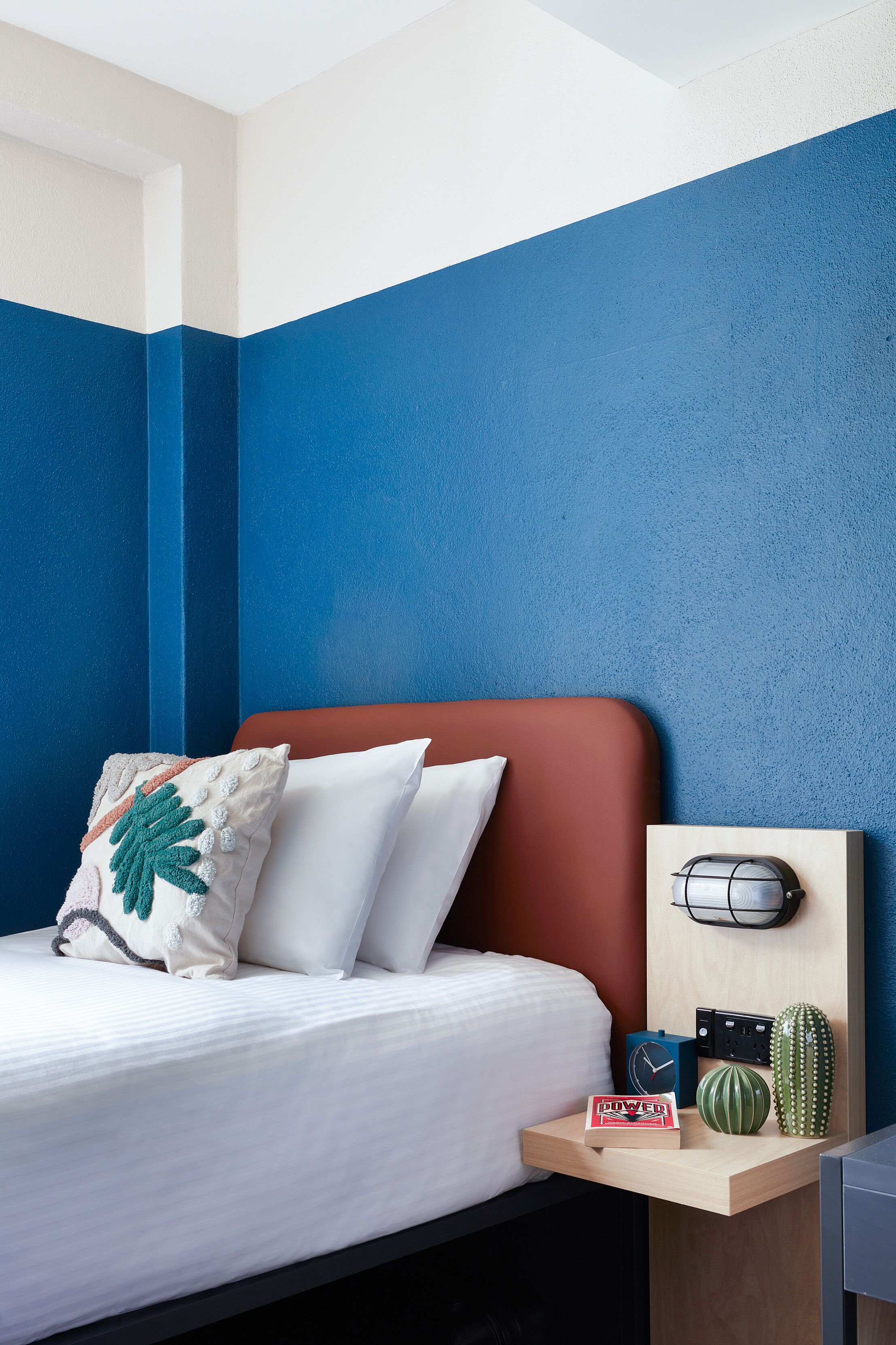 Una cama bien hecha con ropa blanca y un cabecero rojo, una mesita de noche con un reloj, un enchufe y un cactus.
