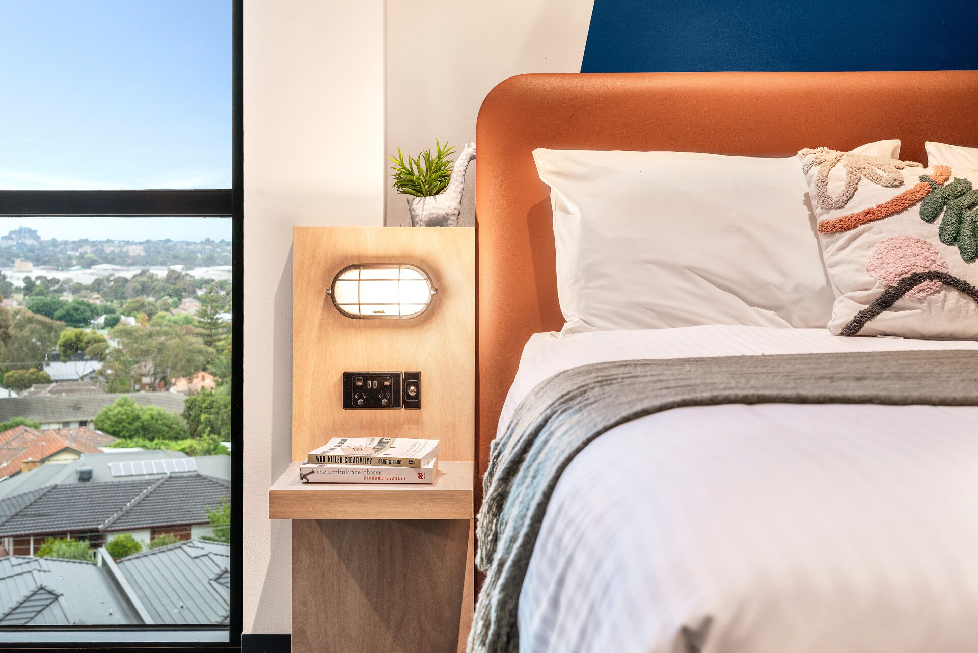 A bedroom with a bed, a nightstand with a lamp, and a view of a cityscape through a window.