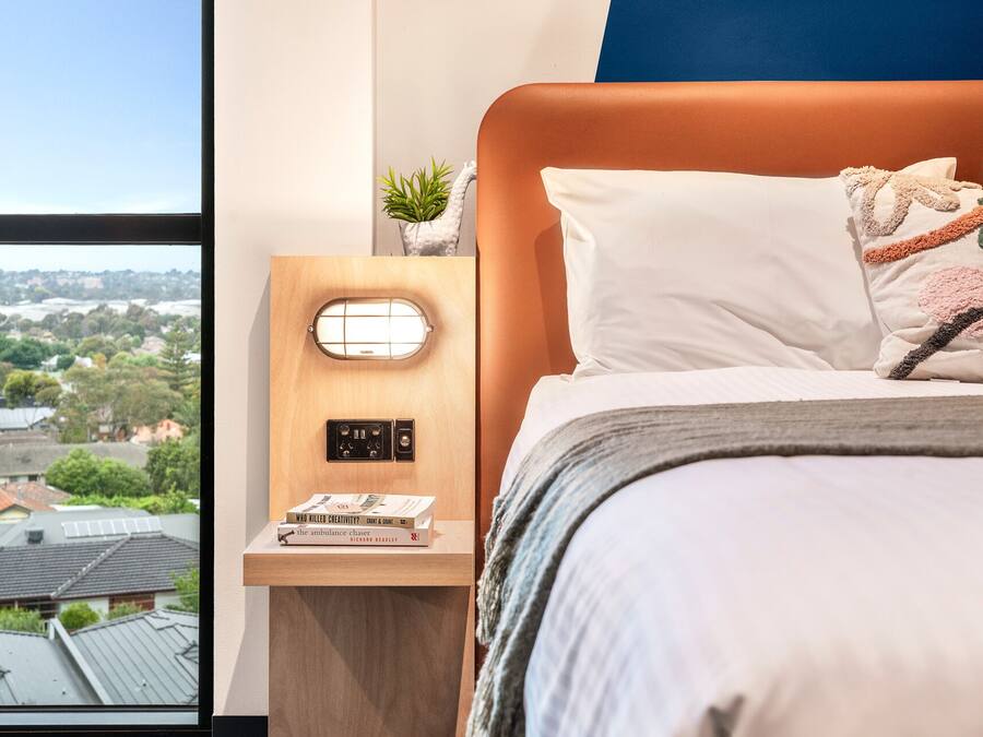 Un dormitorio con cama, una mesita de noche con lámpara y vistas a un paisaje urbano a través de la ventana.