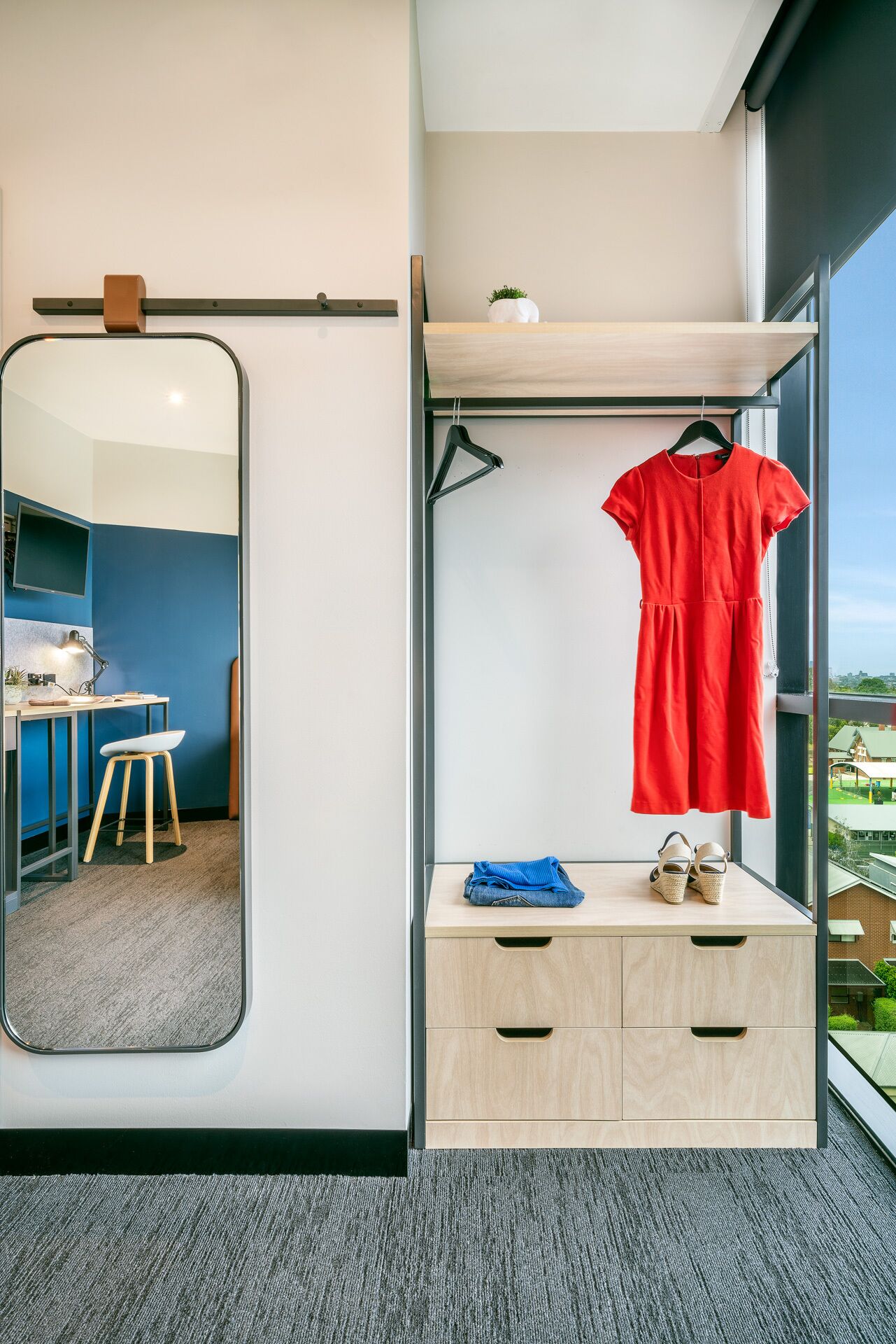A modern room with a large mirror, a desk with a chair, a wardrobe with a red dress, and a view of a green landscape.